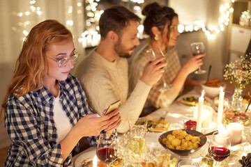 holidays, technology and people concept - young redhead woman in glasses with smartphone at christmas dinner party with friends at home