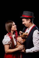 Couple in national ethnic clothing with small dog on hands posing in Studio on black background. Young woman and man or boy and girl in a red and black dress and pet Brussels Griffon in dark room.