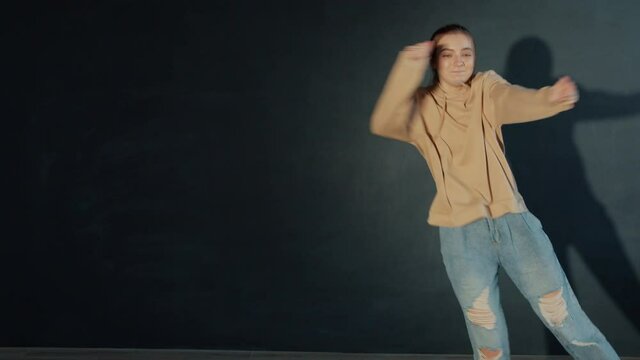 Pretty teen girl is dancing in dark studio performing beautiful creative dance alone having fun, performer is wearing trendy youth clothing. People and hobby concept.