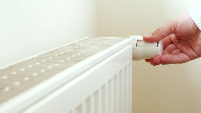 Unrecognizable man's hand setting temperature on central heating system. White metal heating iron household radiator on white wall inside room. Thermostat at home near white wall. Budget in crisis