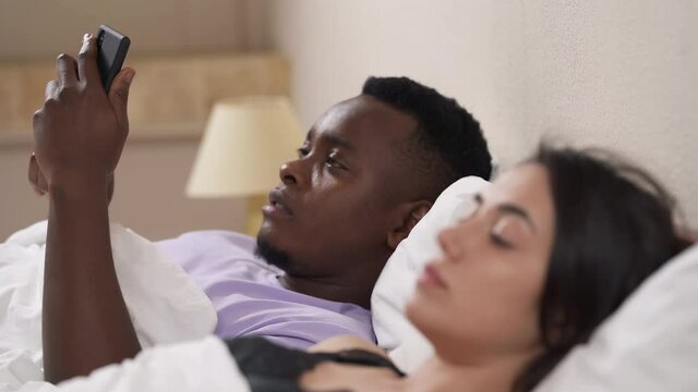 Close-up portrait of interracial couple in bed. African man using mobile phone, offended Caucasian girl turns away from him. Focus changing from guy to girl