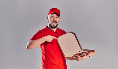 Young bearded delivery man in red uniform shows his finger at pizza box isolated on gray background.