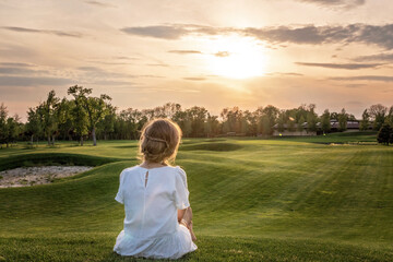A lonely girl in a white dress sitting on golf course looking at sunset sunrise sun. Relaxing and...