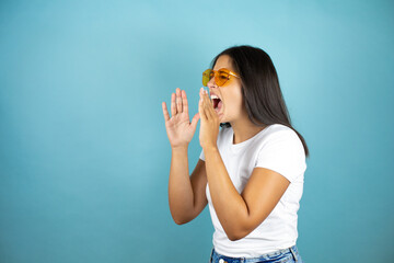 Young beautiful woman wearing sunglasses over isolated blue background shouting and screaming loud to side with hands on mouth