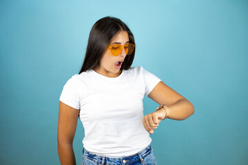 Young beautiful woman wearing sunglasses  standing over isolated blue background surprised and...