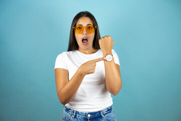Young beautiful woman wearing sunglasses standing over isolated blue background surprised and...