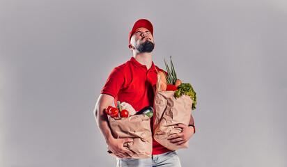 Bearded young man in red T-shirt and cap holding two large heavy bags with food isolated on gray background. Fast food delivery to your home.