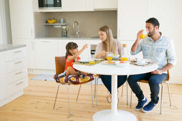 Family couple and girl having breakfast together in kitchen, sitting at dining table, drinking orange juice and talking. Full length, copy space. Family morning concept