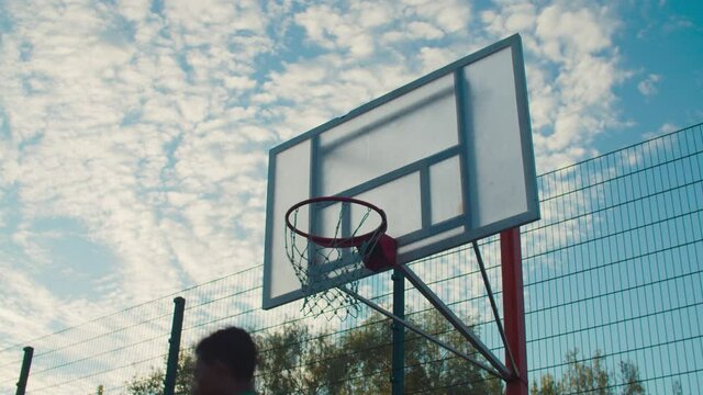 Active athletic african streetball player flying high and dunking with one hand on outdoor basketball court at sunrise. Basketball player performing slam dunk over beautiful morning sky in background.