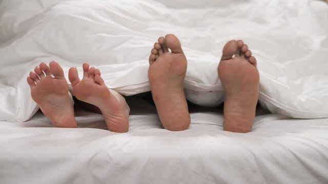 Close-up of bare feet with dancing toes of mixed-race couple, peeking under white blanket. Love and family concept