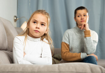 Frustrated small girl sitting at sofa, having conflict with mother at home interior