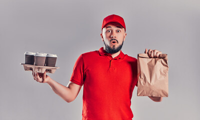 Bearded young man in red T-shirt and cap holds package with fast food and coffee isolated on gray background. Fast delivery from the restaurant to your home.