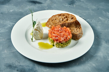 Salmon and avocado tartare, croutons in a white plate on a gray background.