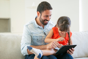 Cheerful girl sitting on her dads lap and using tablet. Father enjoying time with daughter....