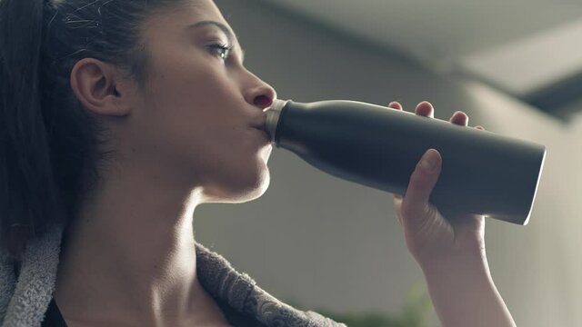 fitness woman drinking from sport bottle during a workout pause at home or gym. Close backlit portrait of active woman drinking from water bottle while resting from training