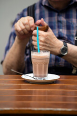 A glass of hot chocolate on a saucer in front of an elderly person who is wearing a check shirt.
