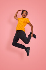 Fototapeta na wymiar Athletic man with dark skin jumps with a happy face in a photo studio near a pink background. Guy in a yellow tshirt, black pants and a sneaker expresses joyful emojis during a photo shoot.