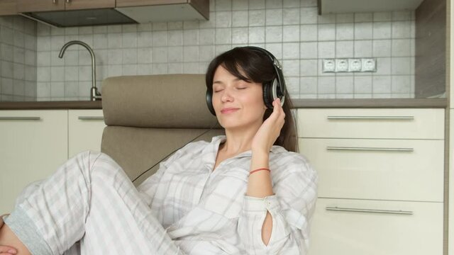 A girl at home sits in a comfortable armchair with her eyes closed and listens to music through headphones. She is dressed in light colored pajamas.