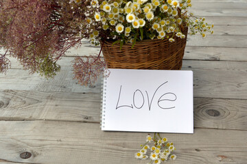 Wicker basket with wild flowers. Text Declaration of love close-up, written on a white sheet. Meadow grasses and daisies in a bag on a wooden background.