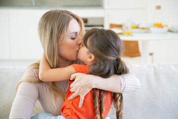 Mom sitting with her little girl on couch, holding kid in arms, kissing and embracing her. Mother...