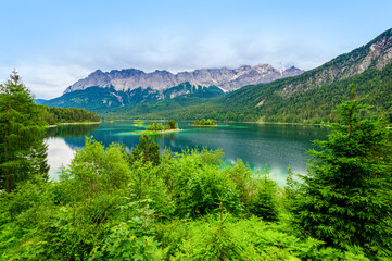 Fototapeta na wymiar Small islands with pine-trees in the middle of Eibsee lake with Zugspitze mountain. Beautiful landscape scenery with paradise beach and clear blue water in German Alps, Bavaria, Germany, Europe.