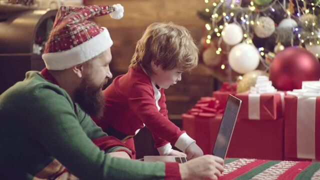 Father and son celebrating Christmas, looking on computer screen near home Christmas interior. Father and son use notebook. Happy people family concept.
