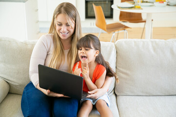 Cute girl sitting near mom and showing open mouth and teeth at computer webcam. Mother and daughter using laptop for video call, talking to grandparents together. Communication concept