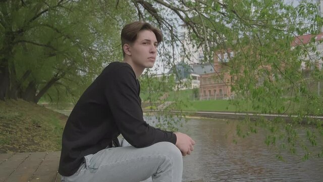The average plan. On a summer day, against the backdrop of the city, a guy sits on a wooden pier in a park near the water. The guy is wearing a black sweater. Looks thoughtfully into the distance