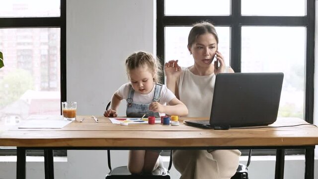 Young mother is talking on phone and having time with her daughter at table in home iroi. Beautiful woman has mobile call while looking at laptop screen and touches cute girl who draws sitting at desk