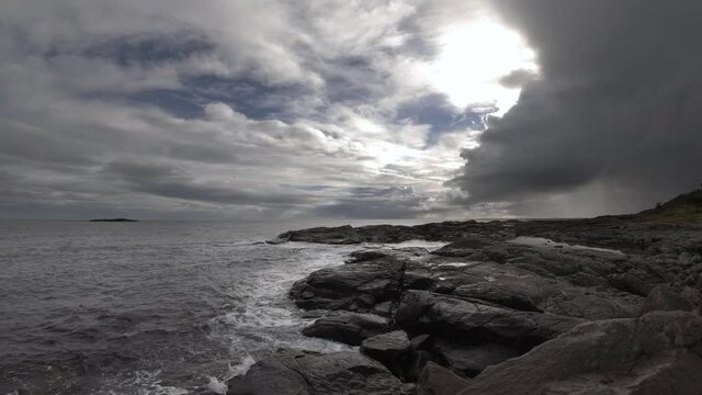 Small waves lapping against black rocks under cloudy dramatic sky in Norway. Rain in the distance.