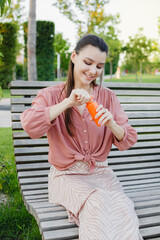 Fototapeta premium Young happy woman opens a bottle with freshly squeezed orange juice. Summer day outdoors in the park. Fresh Orange or carrot juice.