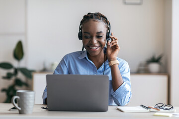 Black female manager wearing headset and using laptop in office, consulting client - obrazy, fototapety, plakaty
