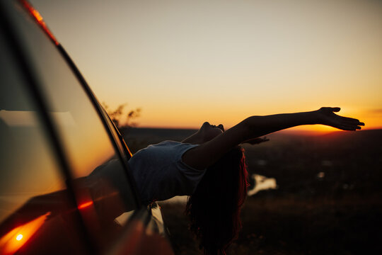 Free young woman with hands up out of a car at amazing sunset. She travels by car catches the wind with her hands from car window. Summer road trip concept. Copy space.