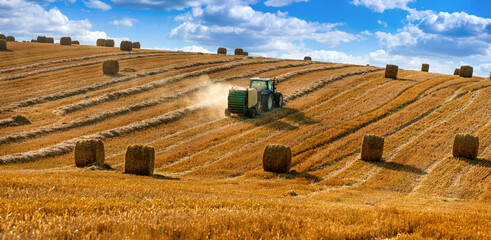 A tractor uses a trailed bale machine to collect straw in the field and make round large bales....