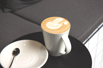 Latte or Cappuccino in a ceramic white cup on a black round table. Morning coffee with foam and a white plate with a spoon. Close up