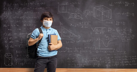 Schoolboy wearing a protective face mask and standing in front of a school chalkboard