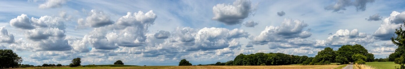 Obraz premium Panorama Himmel mit Wolken über einer Landschaft mit Äckern, Feldern, Bäumen und einem Weg 