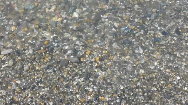 Pure water waves moving on multicolored polished stones and pebbles, closeup view. Sea surf on beach. Sunny summer day. Natural background.