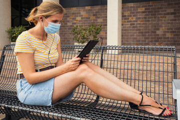 blonde girl in protective mask holding a tablet. A girl sitting on bench in green park with tablet....