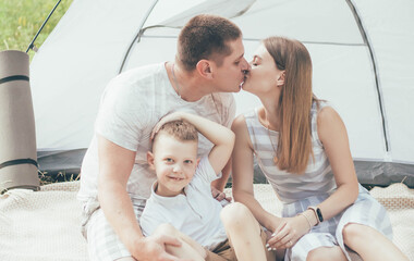 
Family is resting on the river bank with tent dad is kissing mom and the boy is smiling