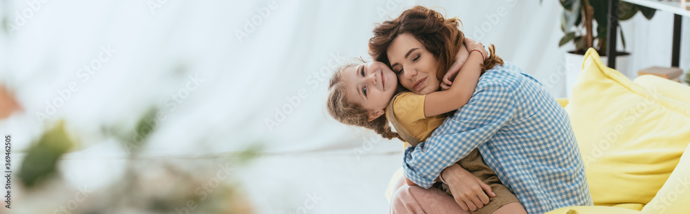 Wall mural horizontal image of happy nanny hugging smiling kid while sitting on sofa