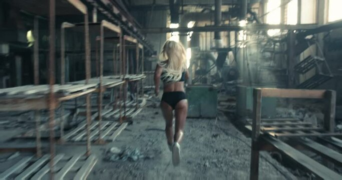 Back view slow motion video of a blonde sportswoman with toned body jogging in a factory early in the morning.