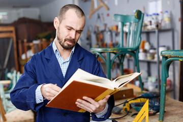 Portrait of positive professional man carpenter standing with book in furniture repair shop. High...