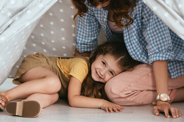 cropped view of babysitter and happy child looking at camera from play tent