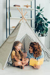 smiling babysitter looking at laughing child while holding book in kids wigwam © LIGHTFIELD STUDIOS