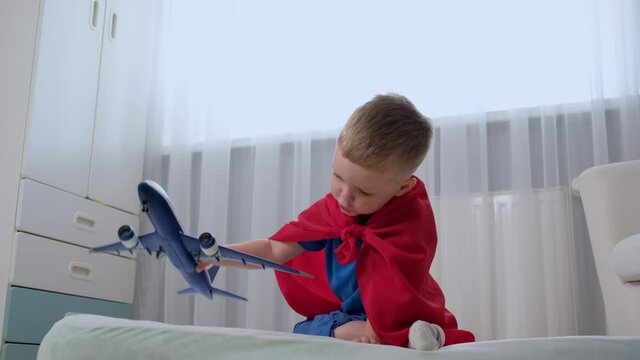 Blond caucasian boy super hero kid is playing with toy air plane in children's room dressed in red raincoat and blue t-shirt. Boy dreams of becoming an air pilot of an airplane. 