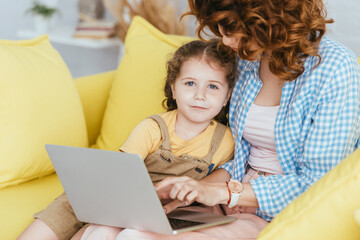 selective focus of adorable child looking at camera near nanny using laptop on sofa