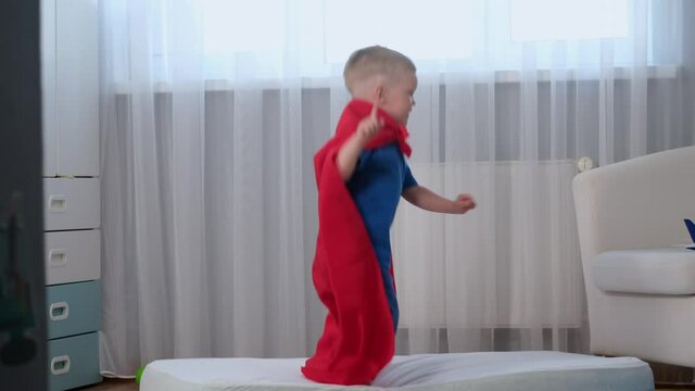Slow motion blond caucasian boy super hero child in red raincoat of hero has fun and jumps in room on mattress like on trampoline up and down falls from trampoline to floor and rises. Travel concept