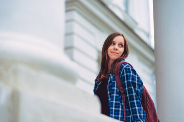Female student outside university building. Low angle of young woman with backpack looking away over shoulder while standing on porch of university building before studies