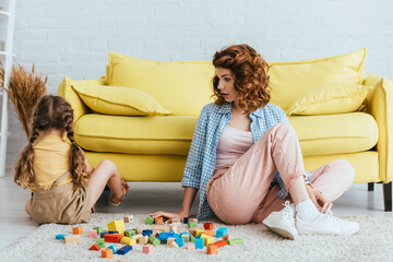 back view of offended child sitting on floor near young nanny and multicolored blocks
